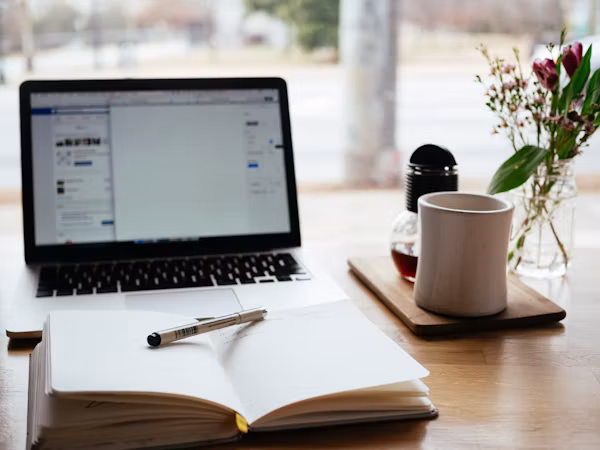 An open notebook and pen beside a laptop, coffee mug, and flowers.